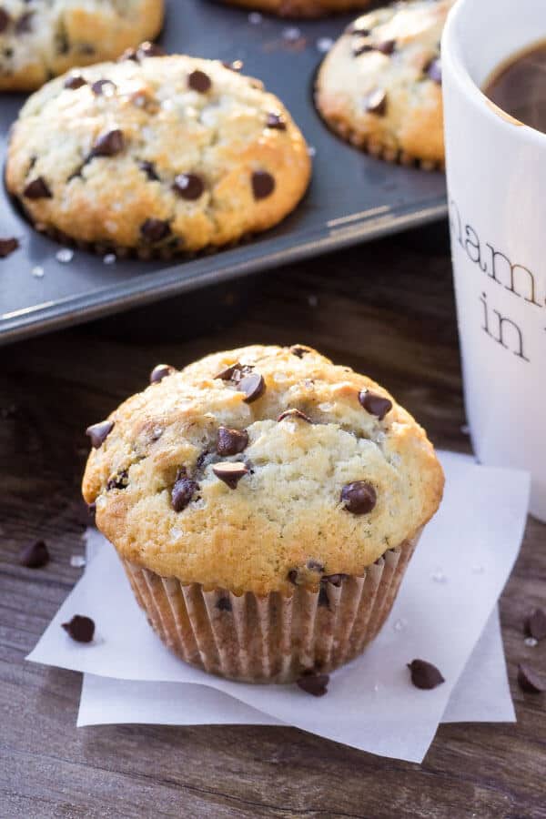 One big, fluffy bakery-style chocolate chip muffin with a cup of coffee and pan full of muffins in the background. 