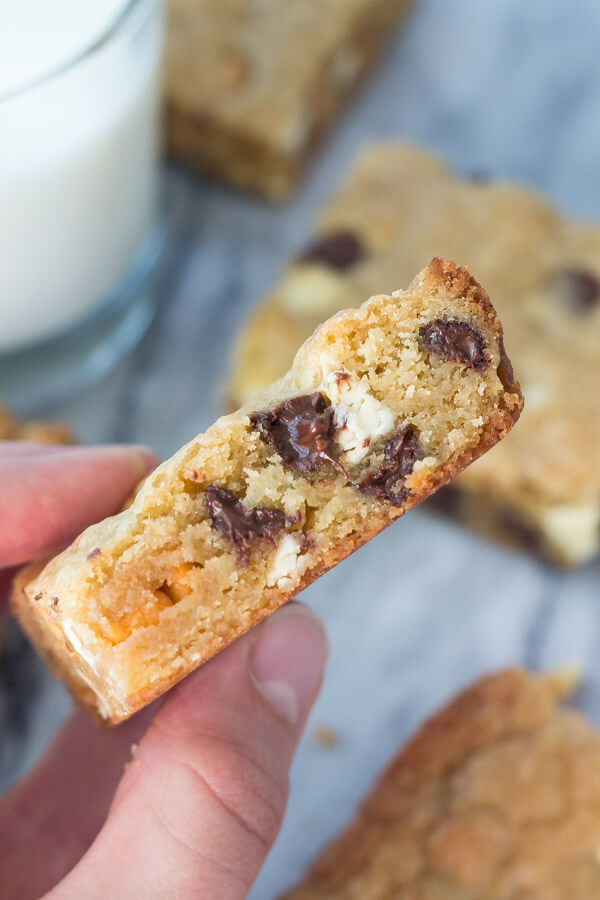 Side shot of a hand holding a triple chip cookie bar. Glass of milk and more cookie bars in the background. 