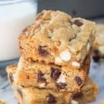 Side shot of 3 stacked triple chip cookie bars with a glass of milk and the cookie bar pan in the background.
