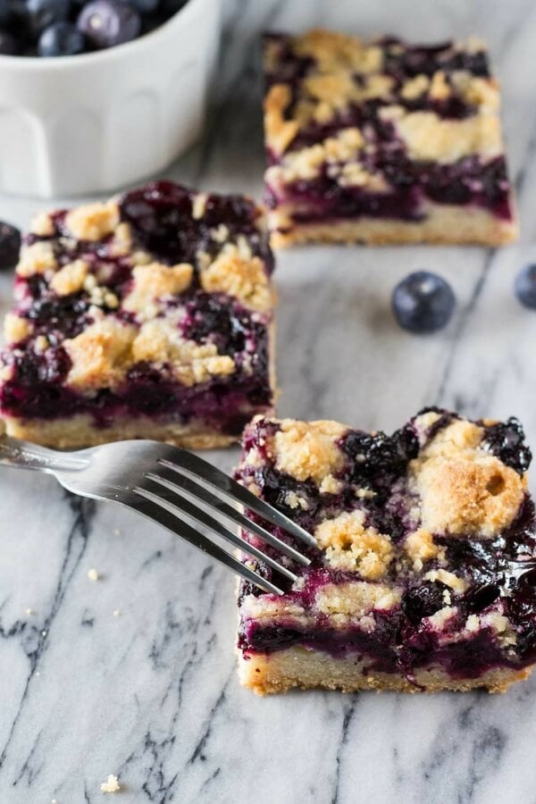 Blueberry crumble bars with a fork about to take a bite of it. A small bowl of blueberries and 2 more bars in the background.