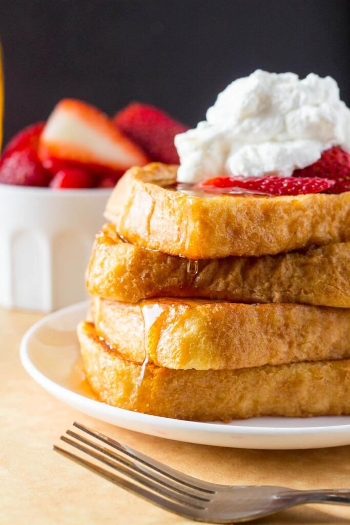 A tall stack for french toast topped with whipped cream and strawberries, then drizzled in maple syrup. 