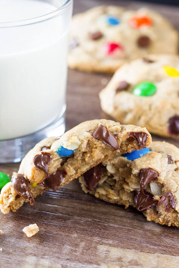 A monster cookie broken in half with melted chocolate chips & M&Ms. Glass of milk in the background. 