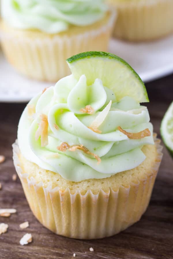 One coconut cupcake with lime frosting, toasted coconut & a slice of lime. Plate of coconut lime cupcakes in the background. 