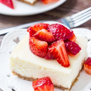 Cheesecake bar topped with chopped strawberries on a white plate. Fork and second cheesecake bar in the background.
