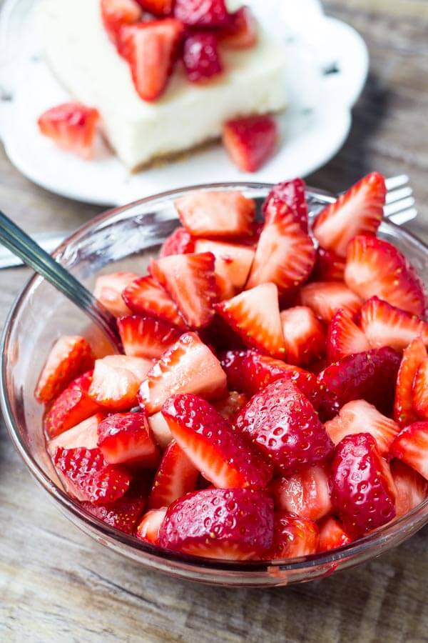 Bowl of chopped sugared strawberries with slice of cheesecake bars in the background. 