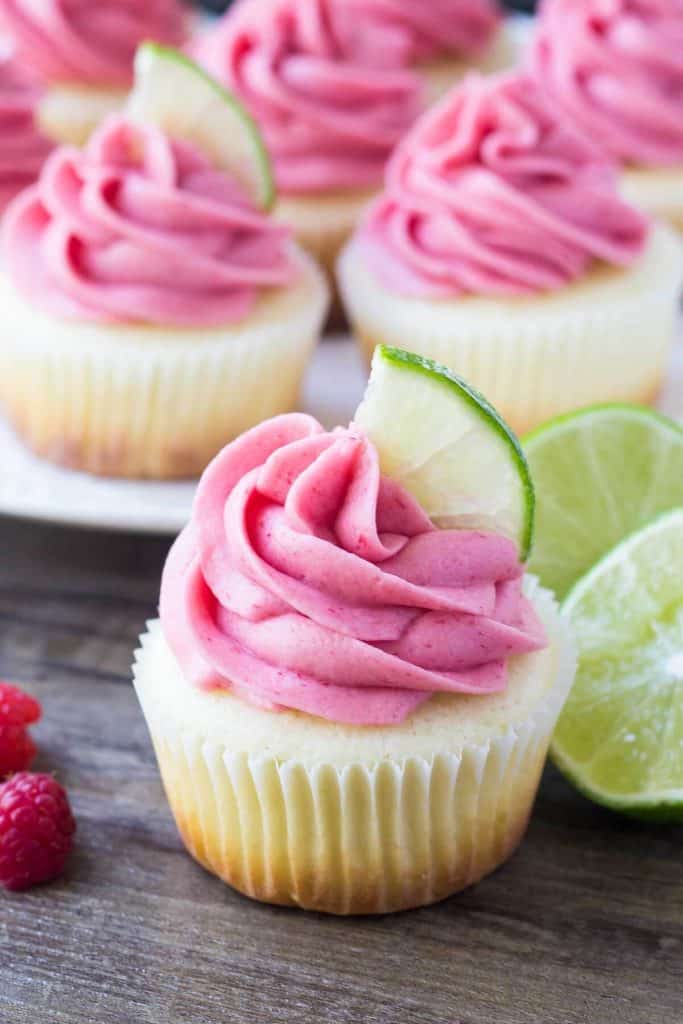 One lime cupcake with raspberry frosting and a plate of cupcakes in the background. Garnished with lime slices and raspberries. 