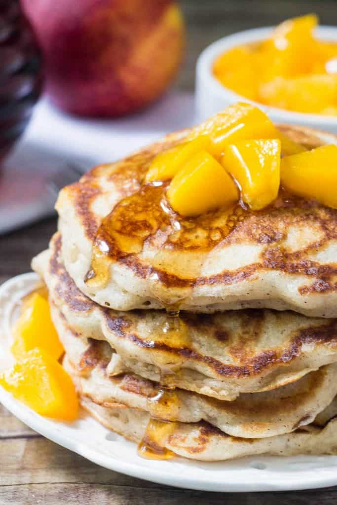 Stack of brown sugar peach pancakes drizzled with syrup and topped with chopped peaches. Jar of syrup of bowl of chopped peaches in the background. 