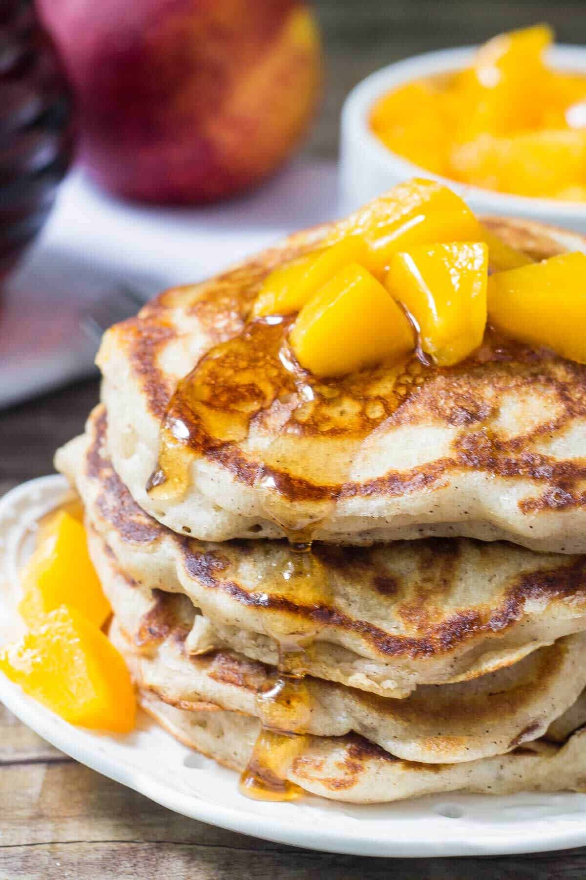 Stack of brown sugar peach pancakes drizzled with syrup and topped with chopped peaches. Jar of syrup of bowl of chopped peaches in the background.