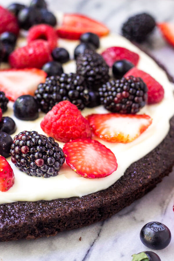 Side angle shot of Fruit Brownie Pizza. A circular brownie, frosted with cream cheese topping and decorated with strawberries, blackberries, raspberries & blueberries.