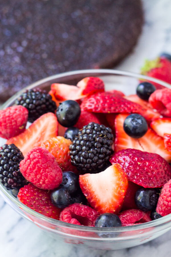 Bowl of strawberries, blackberries, raspberries & blueberries with circular brownie in the background.