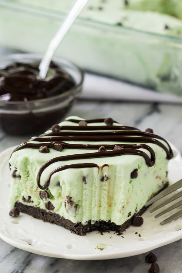 Slice of mint chip ice cream cake with a bite taken out of it. Fork resting on the side of the plate. Bowl of fudge sauce and pan of ice cream cake in the background. 