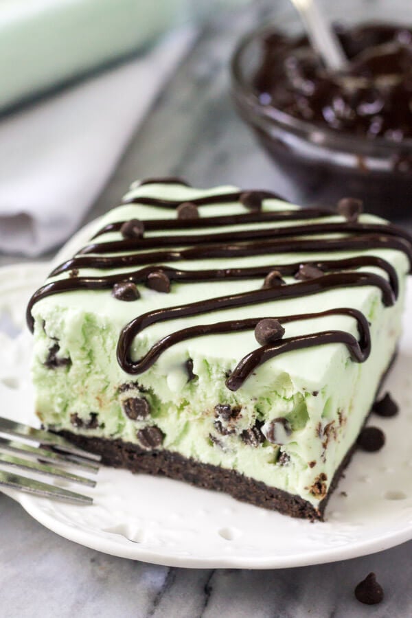Slice of homemade mint chip ice cream cake on a white plate drizzled with chocolate sauce. Bowl of chocolate sauce in the background.
