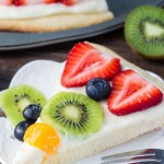 Slice of sugar cookie pizza on a white plate. Full pizza and kiwi in the background.