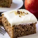 Slice of apple spice cake with cream cheese frosting topped with chopped walnuts. Apple and second slice of cake in the backround.