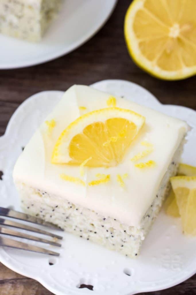 Slice of lemon poppy seed cake with cream cheese frosting on a white plate. Topped with a slice of lemon. ½ lemon and second slice of cake in the background. 