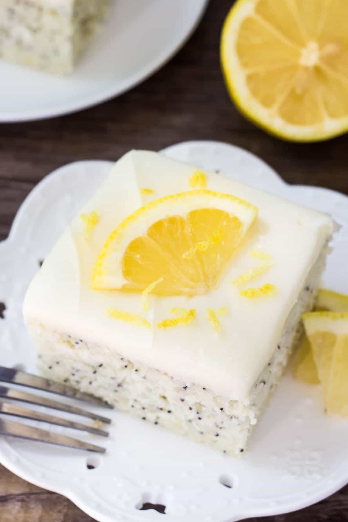 Slice of lemon poppy seed cake with cream cheese frosting on a white plate. Topped with a slice of lemon. 1/2 lemon and second slice of cake in the background.