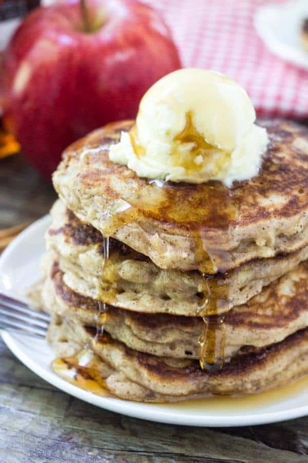 Stack of Apple Pancakes with Brown Sugar and Cinnamon. Topped with whipped butter and drizzled with maple syrup. Apple and bottle of syrup in background. 