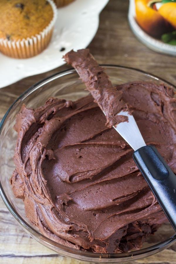 Bowl of chocolate frosting with a plate of pumpkin chocolate chip cupcakes in the background. 