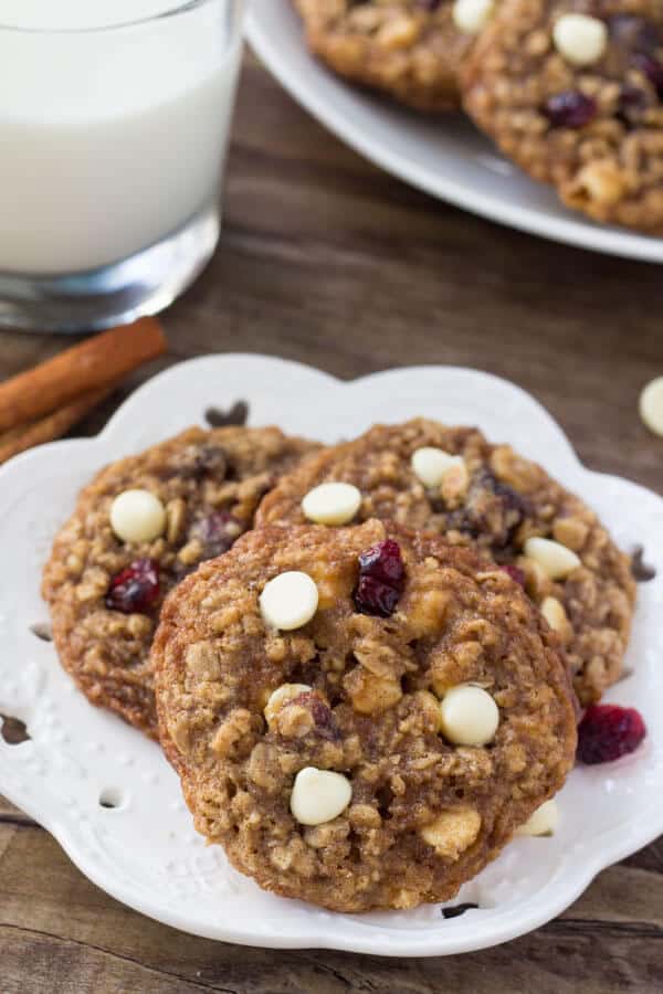 Three white chocolate cranberry oatmeal cookies on a white plate with a glass of milk and a second plate of cookies in the background. 