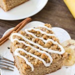 Slice of banana coffee cake with streusel topping and a drizzle of vanilla glaze on a white plate. Second piece of cake and bunch of bananas in the background.