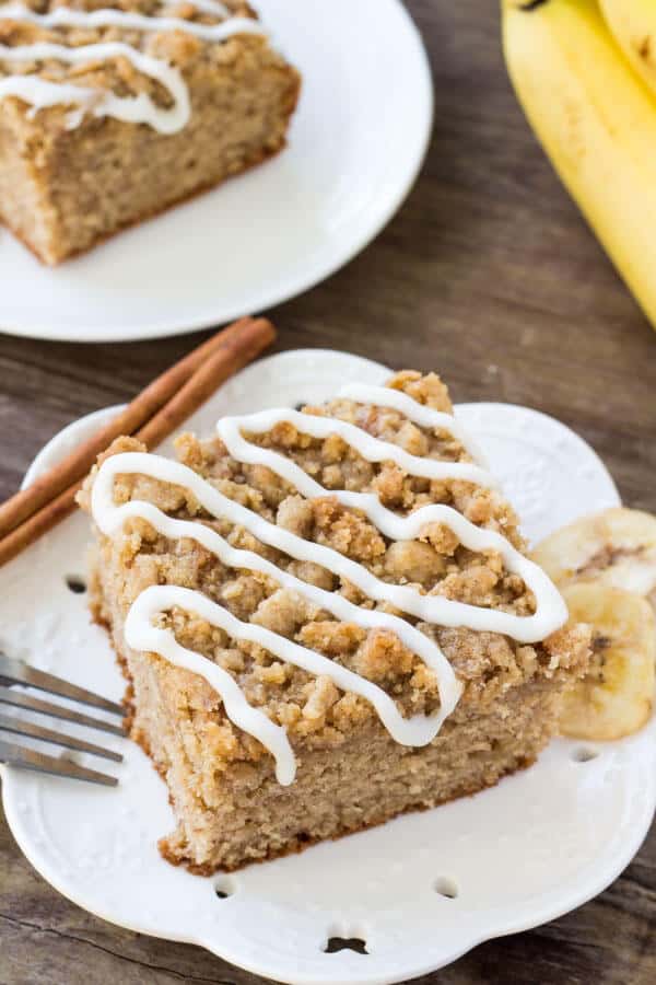 Slice of banana coffee cake with streusel topping and a drizzle of vanilla glaze on a white plate. Second piece of cake and bunch of bananas in the background.