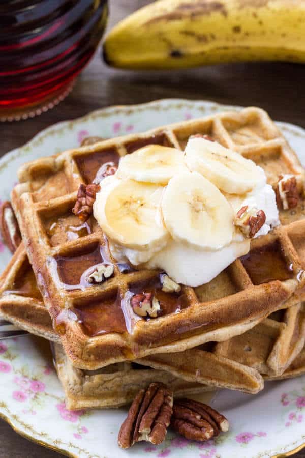 A stack of fluffy banana waffles taste like banana bread topped with maple syrup, whipped cream & sliced bananas. 