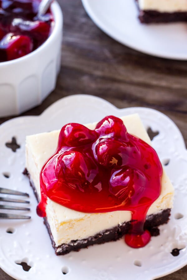 Overhead view of a slice of brownie cheesecake bars topped with cherries. With a bowl of cherry topping in the background. 