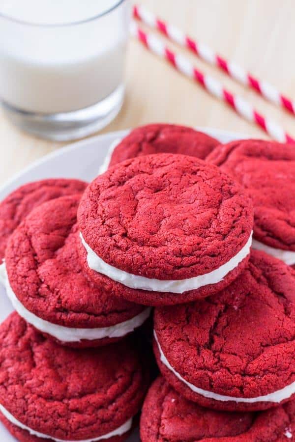 A plate of red velvet sandwich cookies with cream cheese frosting and a glass of milk. 