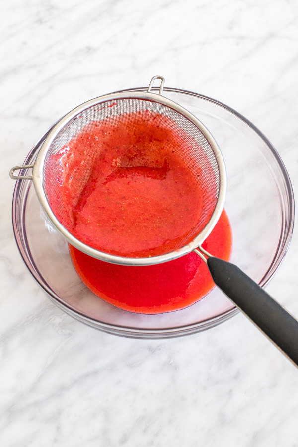 Strawberry puree being pushed through a metal sieve into a bowl.