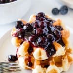 A stack of 2 small waffles topped with blueberry syrup with a bowl of blueberry sauce in the background.