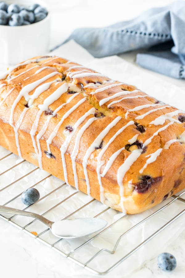 Blueberry Bread drizzled with vanilla glaze on a cooling rack with a small bowl of blueberries in the background. 