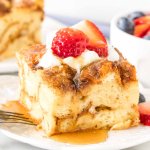 Slice of french toast bake with whipped cream, strawberries and maple syrup on top on a plate.