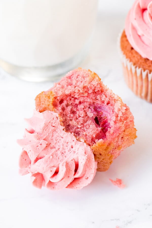 A strawberry cupcake with strawberry chunks in the batter with a bite taken out of it. 