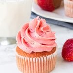 A strawberry cupcake with strawberry frosting with a glass of milk and plate of strawberry cupcakes in the background.