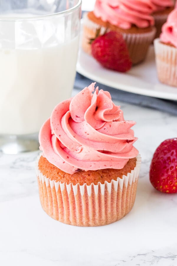 A strawberry cupcake with strawberry frosting with a glass of milk and plate of strawberry cupcakes in the background. 