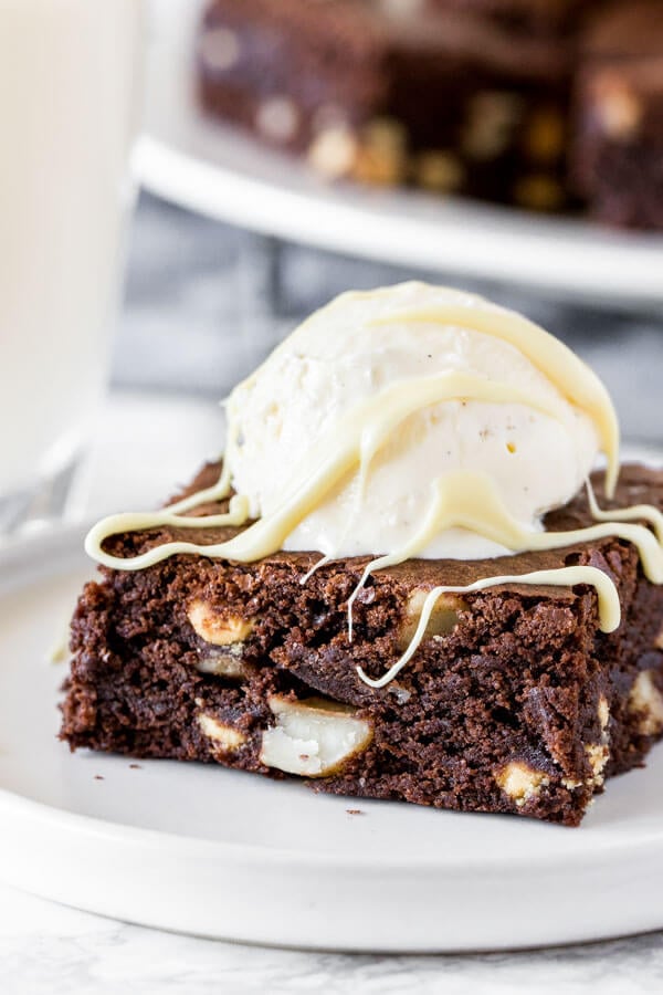 A fudgy white chocolate macadamia nut brownie on a white plate with a scoop of vanilla ice cream to make a brownie sundae. 