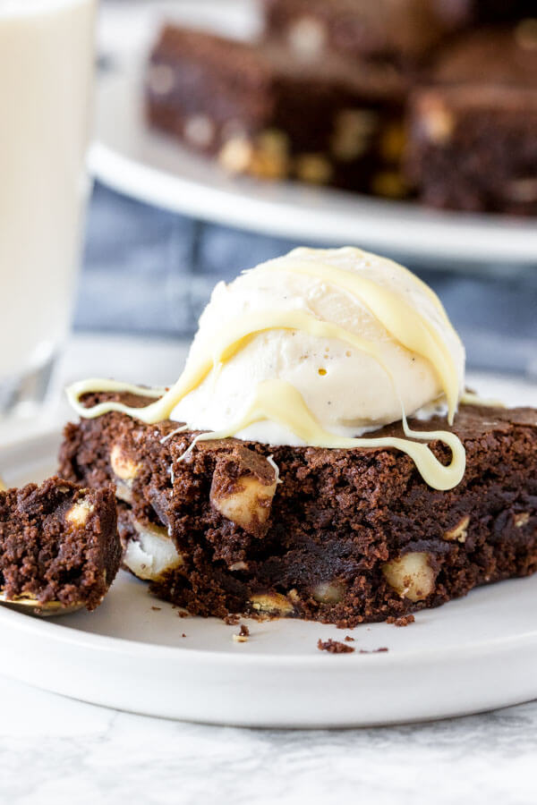 A fudgy white chocolate macadamia brownie with a scoop of vanilla ice cream and melted white chocolate drizzle with a plate of brownies in the background. 