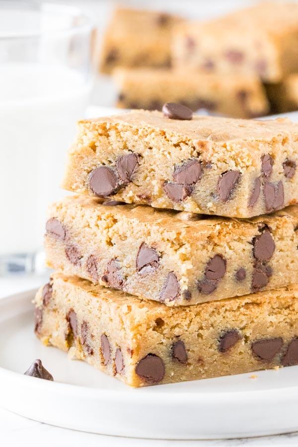 A stack of 3 chocolate chip blondies on a plate with a glass of milk and large plate of blondies in the background. 