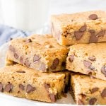 Chewy chocolate chip blondies cut into squares and stacked on a white plate with a glass of milk in the background.