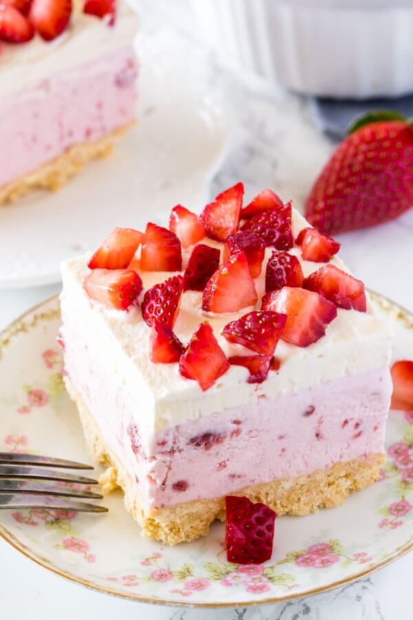 A square slice of strawberry shortcake ice cream cake on a white plate topped with whipped cream and pieces of fresh berries. 