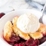 A bowl of fresh cherry cobbler with a flaky biscuit topping and scoop of vanilla ice cream on top.