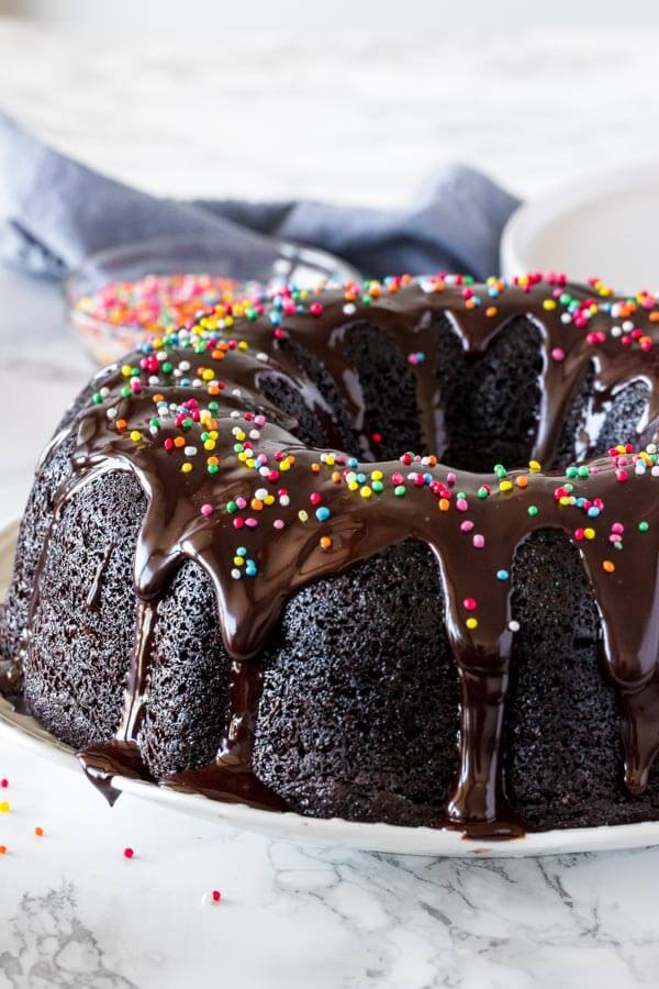 A chocolate bundt cake on a white plate that's been drizzled with chocolate ganache and topped with sprinkles.
