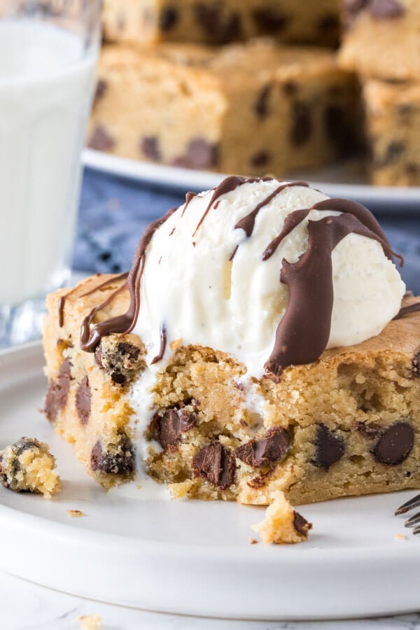 A chocolate chip cookie bars with a scoop of ice cream on top and a drizzle of chocolate on top with a bite taken out of it. 