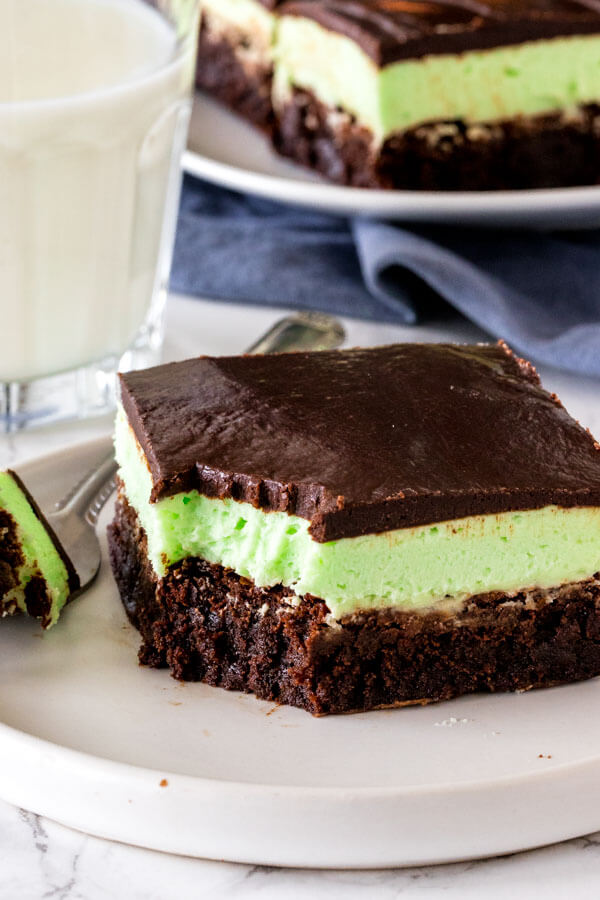 A layered mint brownies with a bite taken out of it on a plate with a glass of milk to show the fudginess of the brownie and creaminess of the mint filling. 