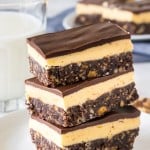A stack of 3 classic Nanaimo bars on a white plate with a glass of milk in the background.