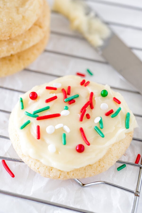 A round sugar cookie with white frosting and Christmas sprinkles on a cooling rack.