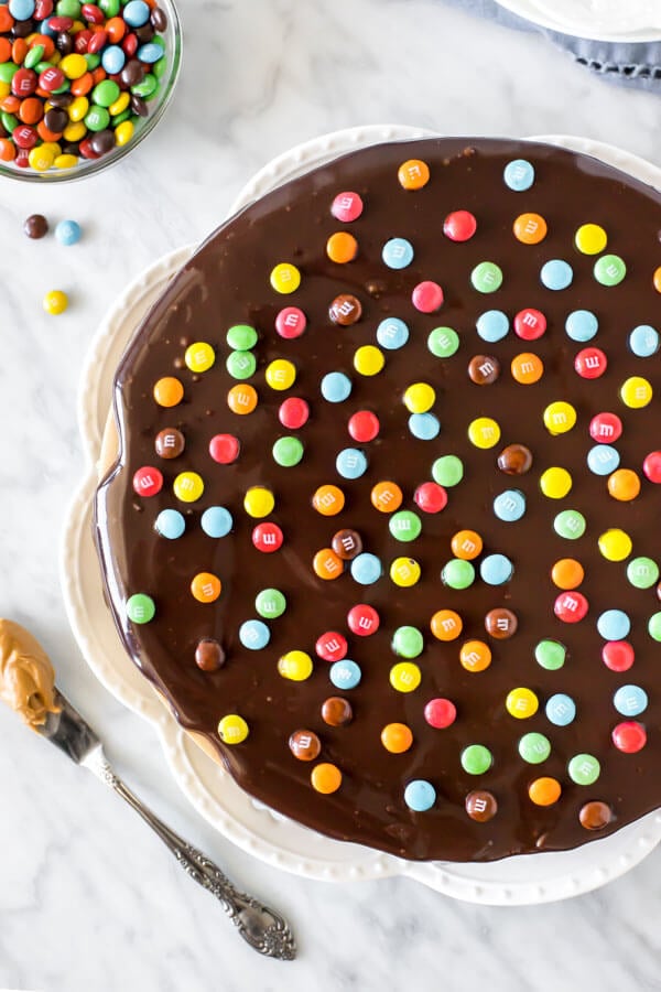 Cheesecake topped with chocolate ganache and M&M candies on a white plate, shot from above. 