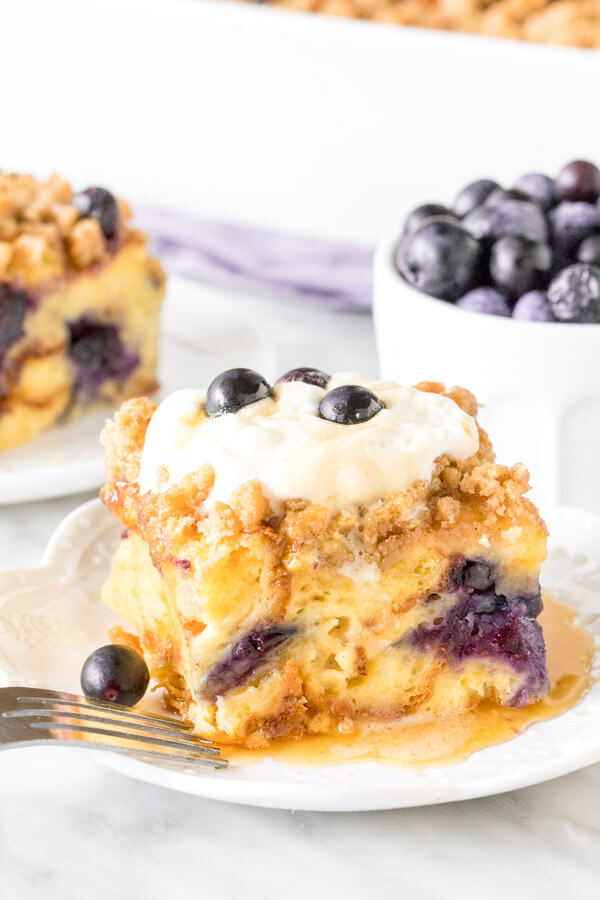 Slice of blueberry French toast casserole with whipped cream on top and drizzled in syrup. Bowl of blueberries and second piece in the background. 