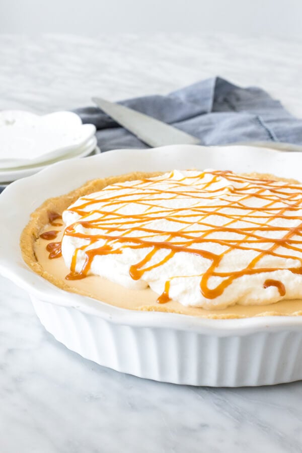 No-Bake caramel cream pie in a white pie plate topped with whipped cream and a drizzle of caramel sauce. 