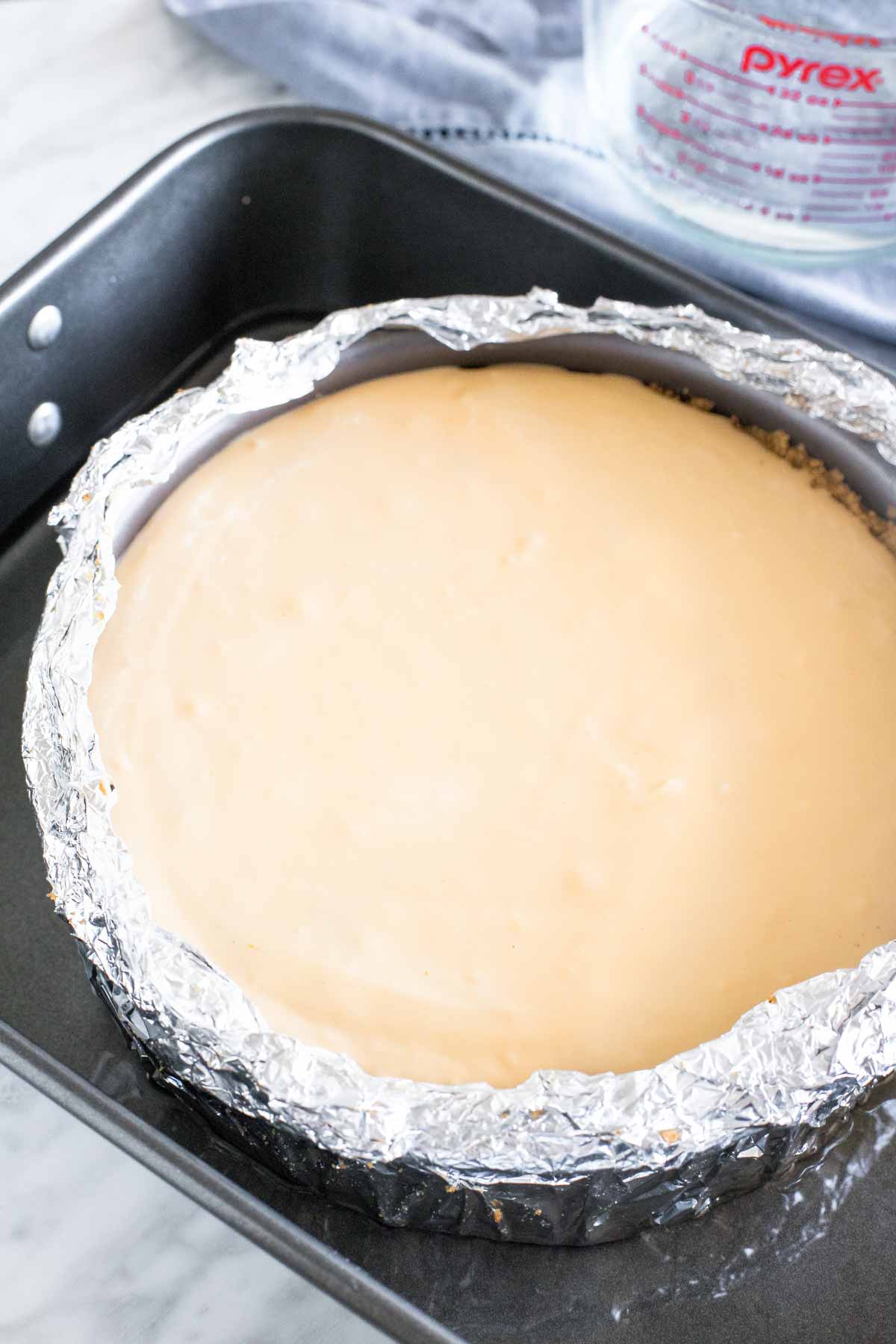 Unbaked cheesecake in a springform pan wrapped in tinfoil in a large roasting pan. 